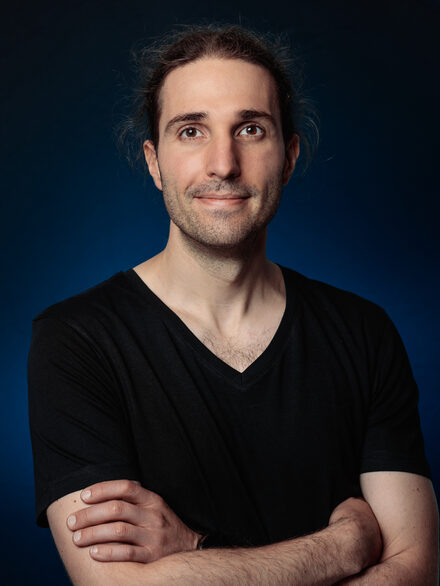 Portrait of Johannes Otterbach in a black T-shirt, arms crossed, against a dark blue background
